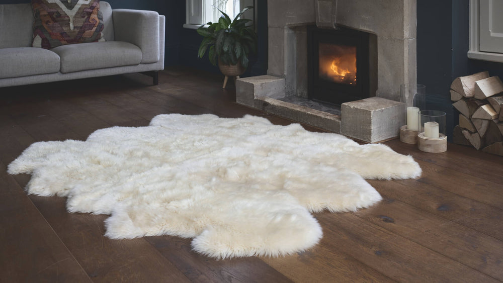 An extra large ivory sheepskin rug laid out on a wooden floor in a living room setting with a fireplace and furniture in the background.