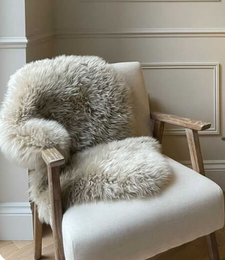 A light beige/cream colored sheepskin rug with a delicate iridescent silver undertone, displayed on the floor under a wooden chair with a white cushion, next to a small round table with a vase of flowers.