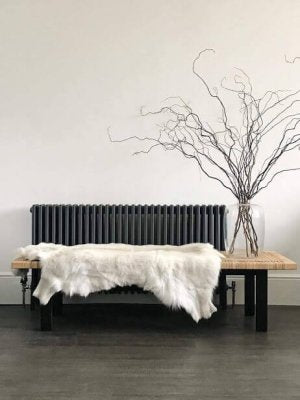Black radiator with a wooden bench and white fur reindeer  hide against a plain wall.