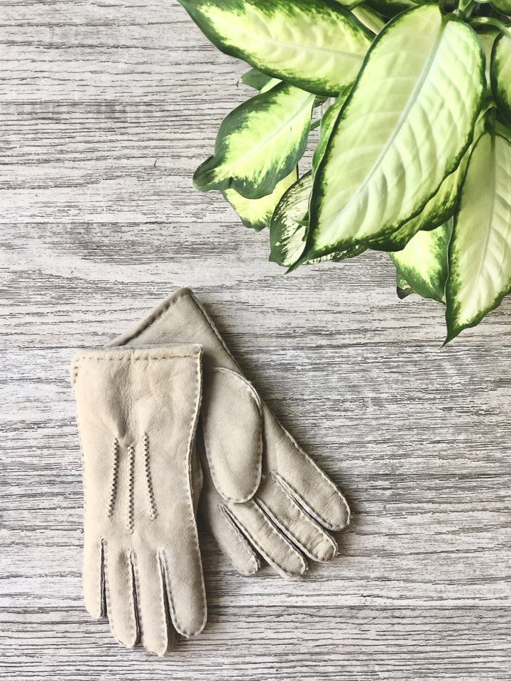 A pair of beige ladies sheepskin gloves laid flat on a wooden surface, with a green leaf partially visible in the background.