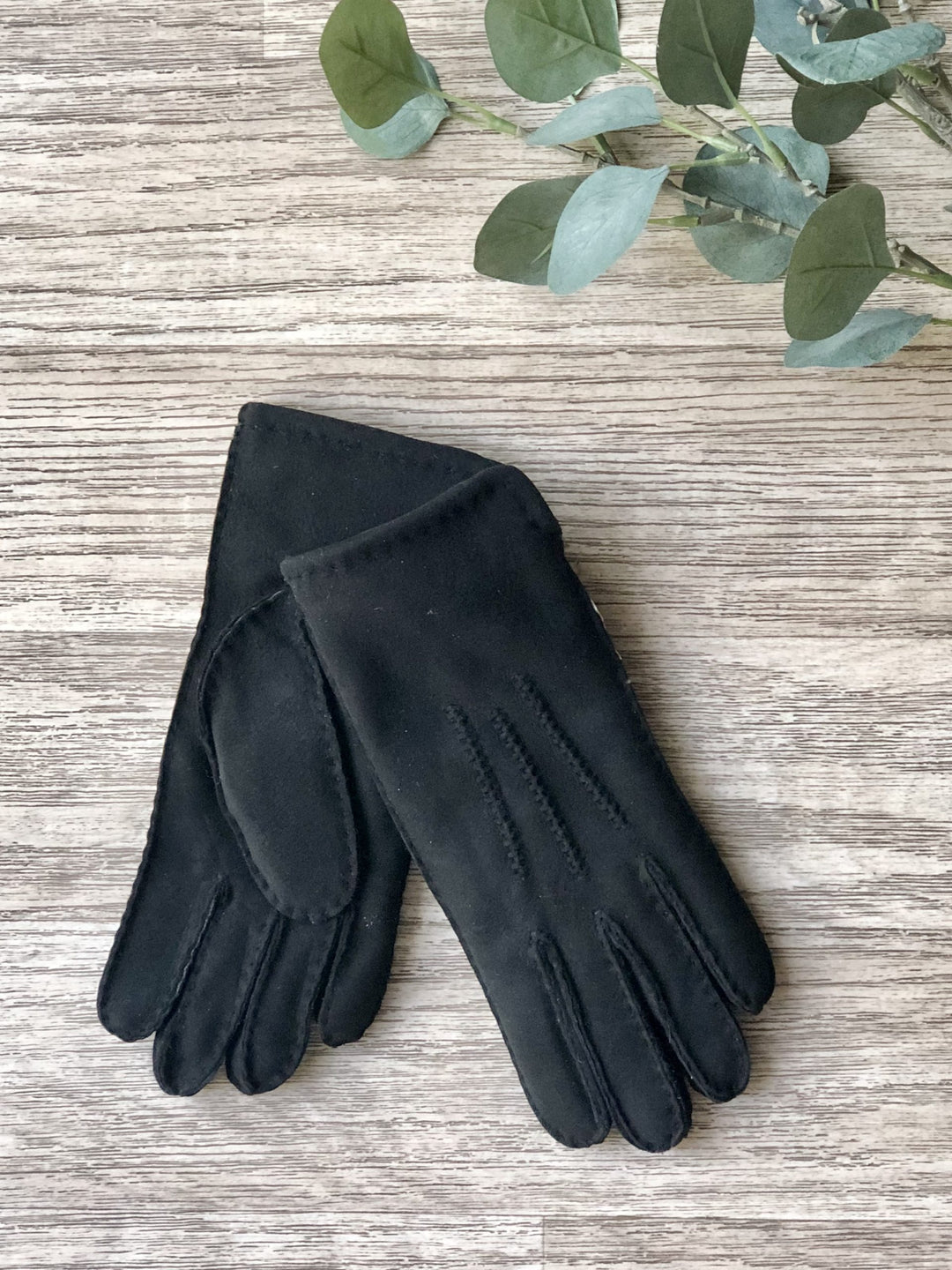A pair of ladies black sheepskin gloves laid flat on a wooden surface, with a green leaf partially visible in the background.