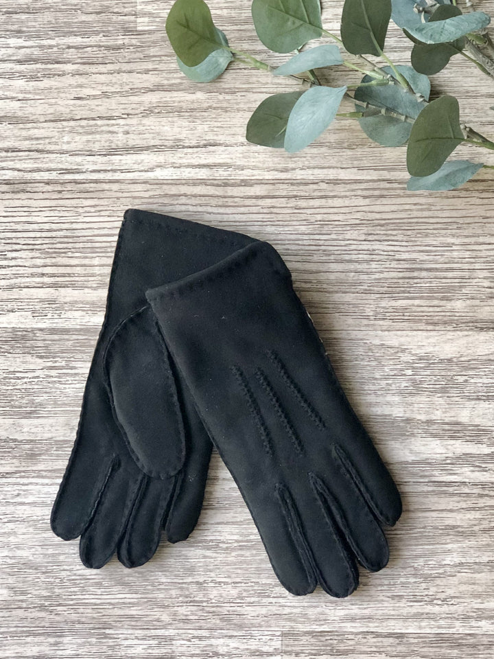 A pair of ladies black sheepskin gloves laid flat on a wooden surface, with a green leaf partially visible in the background.
