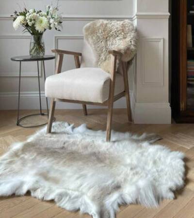 A light beige/cream colored sheepskin rug with a delicate iridescent silver undertone, displayed on the floor under a wooden chair with a white cushion, next to a small round table with a vase of flowers.