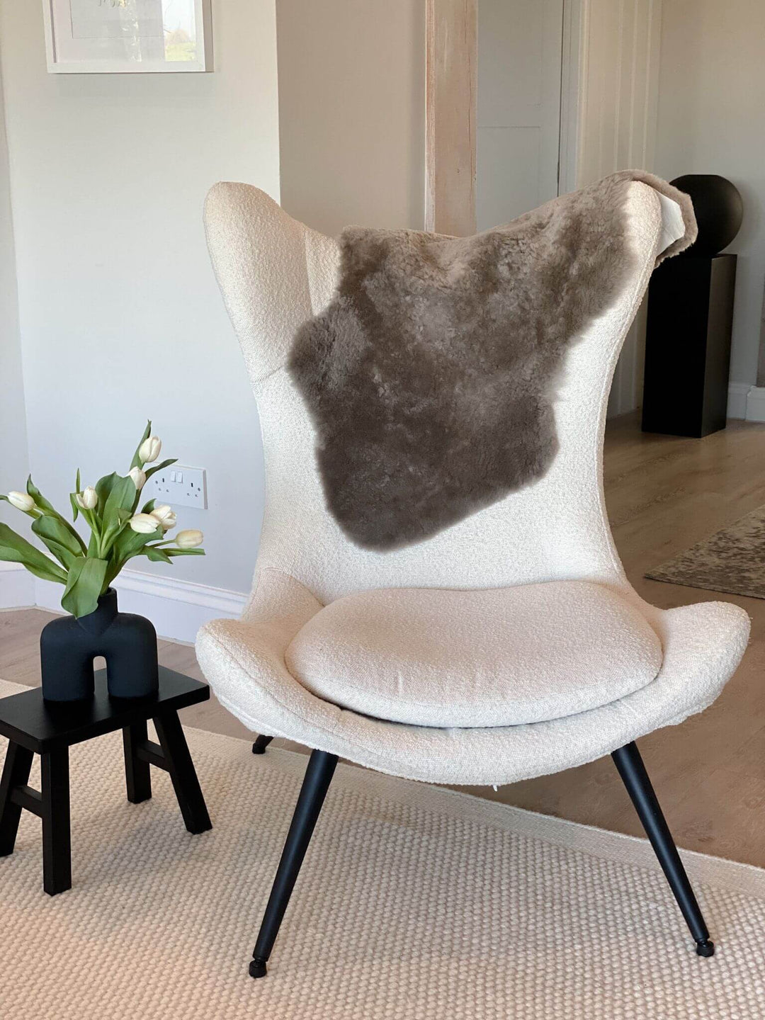 A short wool sheepskin rug in a taupe color, placed on a chair, with a plant pot on a side table in the background.